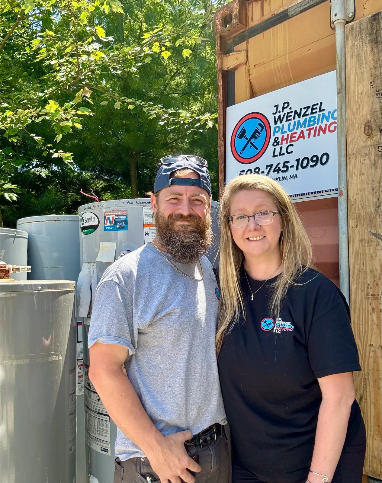 A man and woman standing outdoors in front of a sign for J.P. Wenzel Plumbing & Heating LLC, smiling at the camera. The man has a beard, is wearing a gray t-shirt, baseball cap backwards, and sunglasses on his head. The woman has long blonde hair, glasses, and is wearing a black t-shirt with company branding.