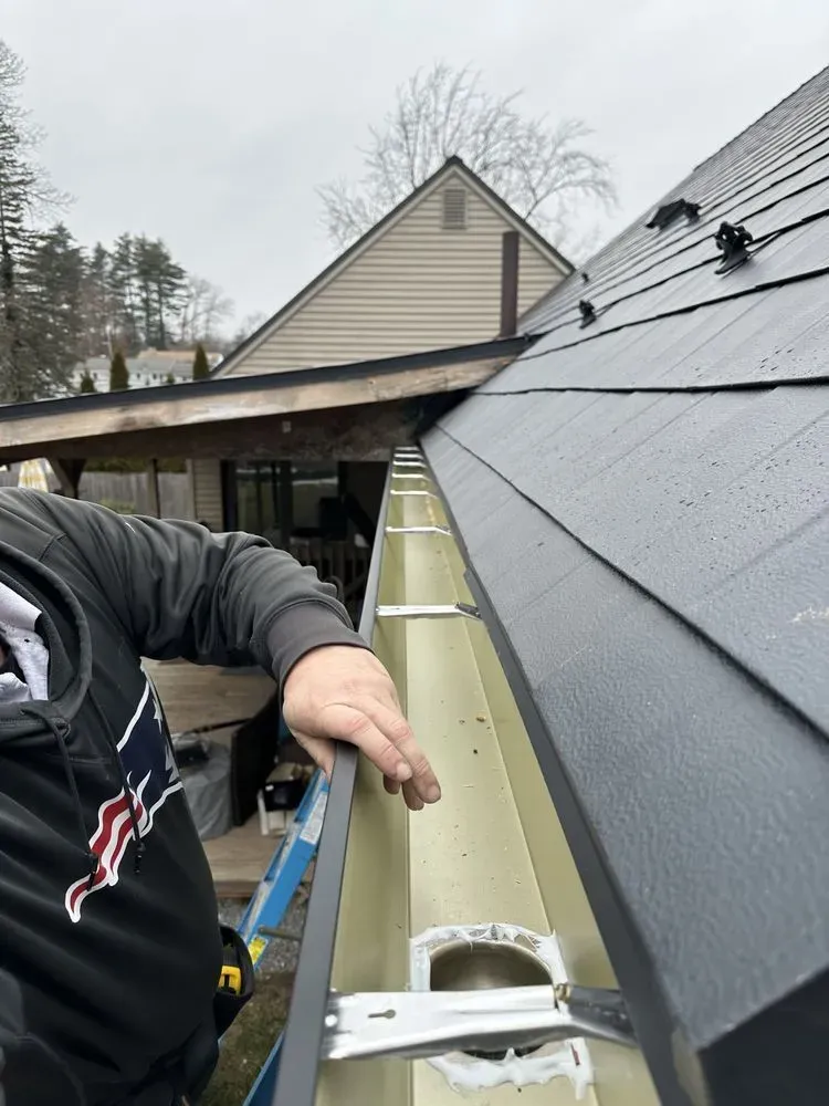 Worker inspecting rain gutter beside a black-roofed house with ladder and tools