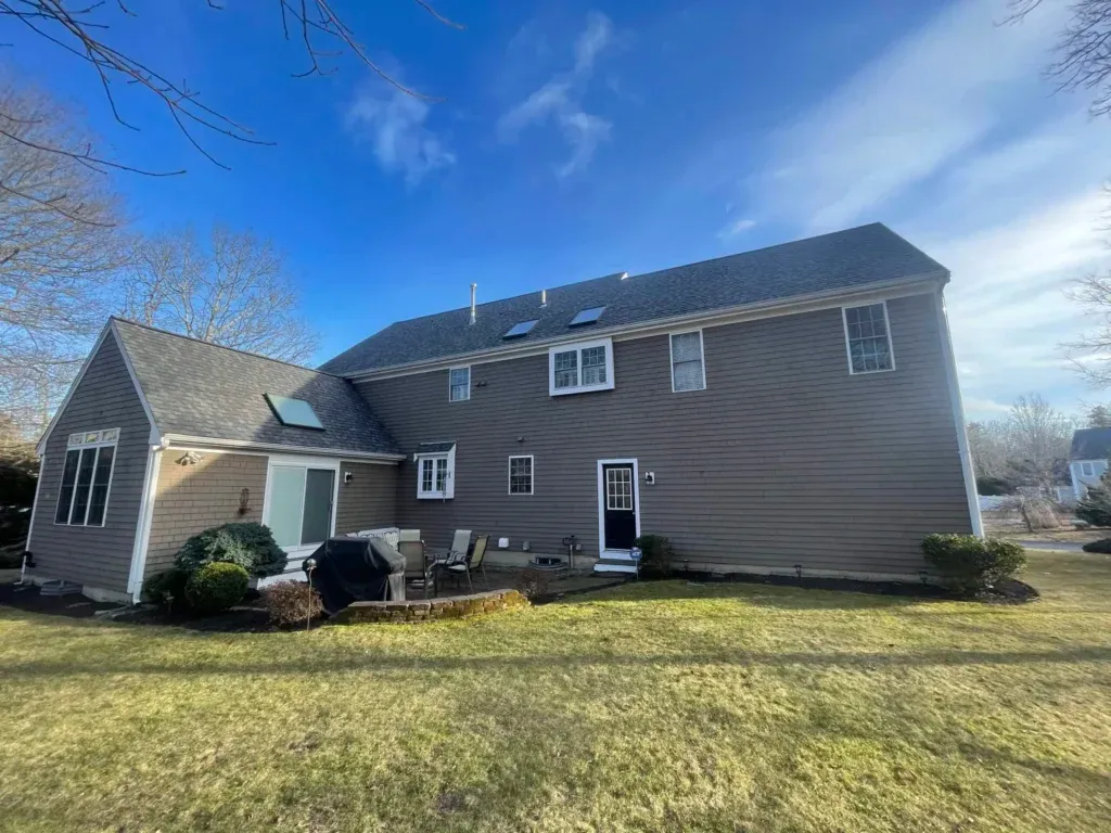 A large, two-story house with beige siding and multiple windows. There is a patio in the backyard with outdoor furniture and a grill. The yard is well-maintained with some shrubs and trees. The sky is clear with a few clouds.