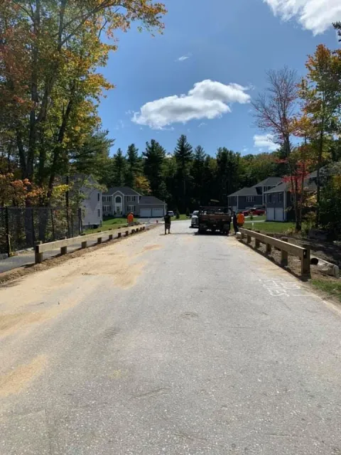 A construction crew works on a paved residential street lined with new wooden guardrails under a bright, sunny sky.