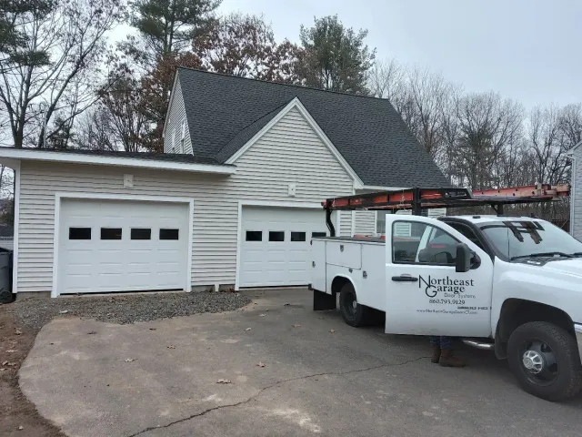 White service truck parked beside a two-car garage with a dark shingled roof and bare trees behind it
