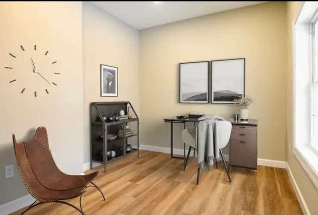 A cozy home office with a wooden desk, two chairs, and wall art. Features a large window, a wall clock, and a shelving unit with decorative items. The room has light yellow walls and hardwood flooring.