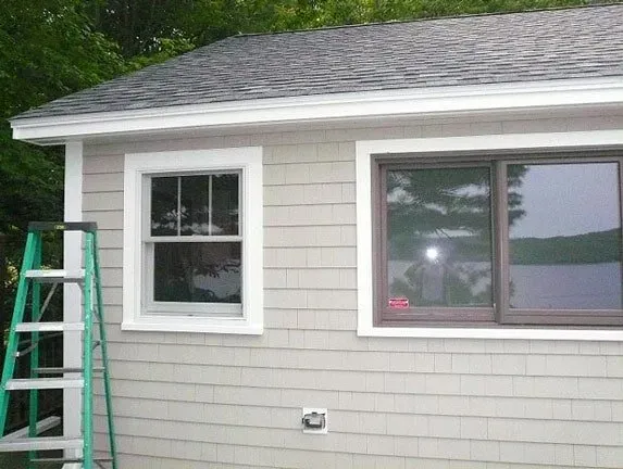 A green ladder stands against the side of a light gray shingled house with two windows and a roof overhang.