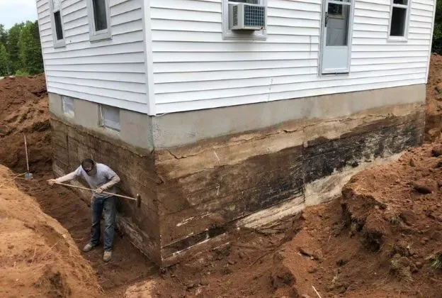 Person digging around a house foundation, showing exposed earth layers. White siding, air conditioner.