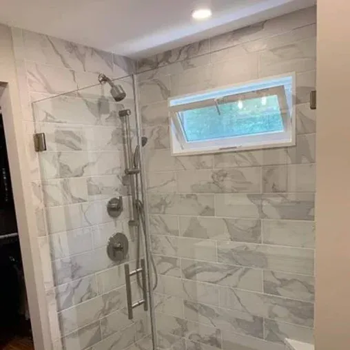 A modern shower with marble-patterned tiles, a glass door, multiple showerheads, and a small rectangular window positioned vertically, opening sideways.