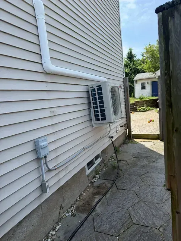 A mini-split air conditioner unit is mounted on the light-colored vinyl siding of a house exterior near a stone path.