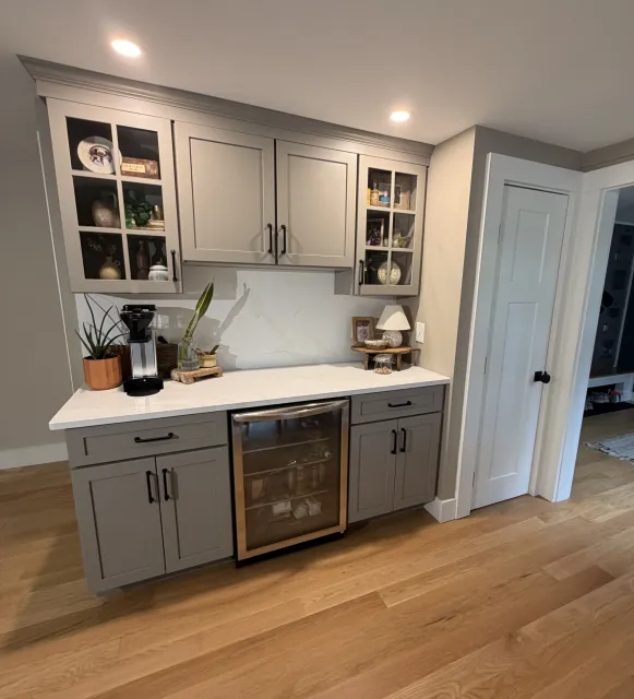 Gray kitchen cabinets with glass doors displaying decorative items, a white marble countertop, and a rustic wooden table below with plants and small decor items in front of the cabinets.