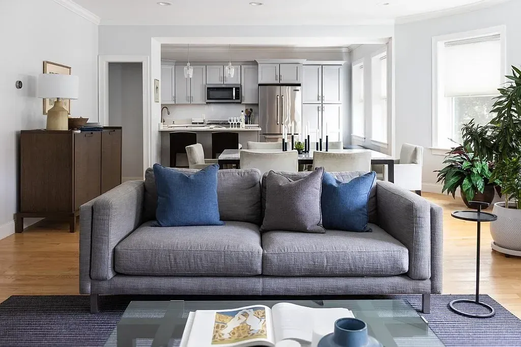 Living room with a gray sofa, blue and gray pillows, a glass coffee table, and a kitchen and dining area in the background with white cabinets and large windows.