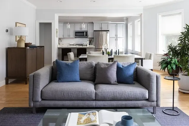 Living room with a gray sofa, blue and gray pillows, a glass coffee table, and a kitchen and dining area in the background with white cabinets and large windows.