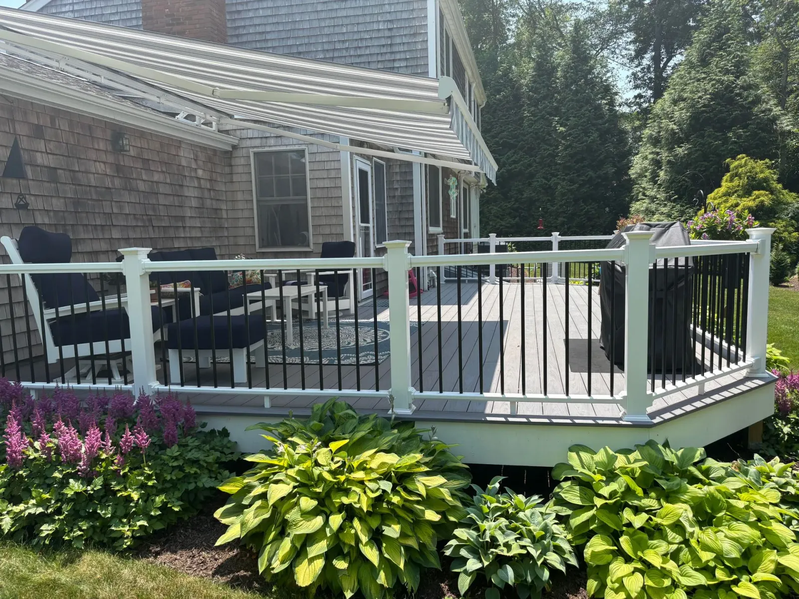 Deck with white railing and black spindles, attached to a house with a pergola. Lush green foliage surrounds the deck.