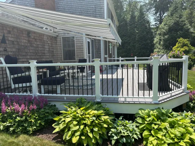 Deck with white railing and black spindles, attached to a house with a pergola. Lush green foliage surrounds the deck.