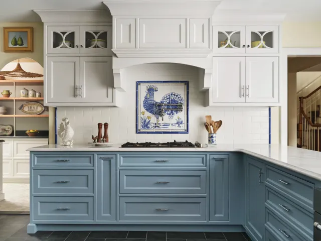 Kitchen with white upper cabinets and blue lower cabinets, decorative tile with a rooster on the wall, and a white countertop.