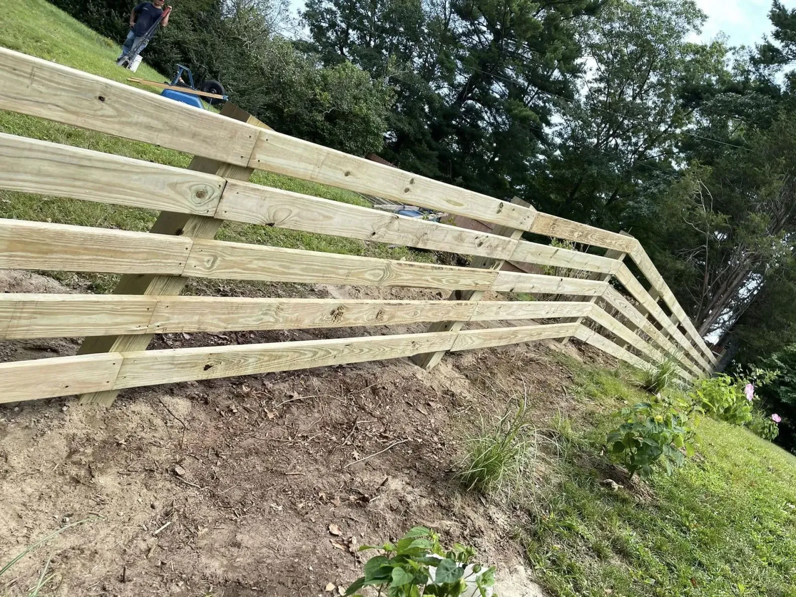 Wooden fence built on a hillside. Person in background, green grass and trees.