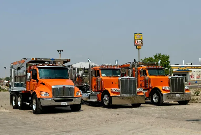 Three orange semi trucks parked side by side in a lot under a clear sky