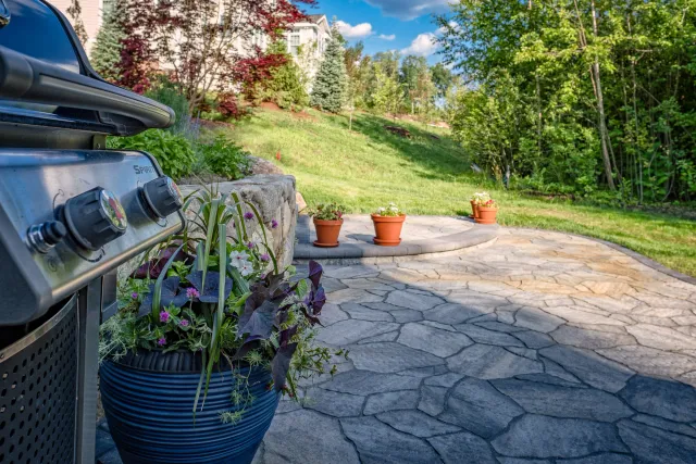 Patio with stone flooring, a grill on the left, potted plants and flowers, lawn, and trees in background under a blue sky.