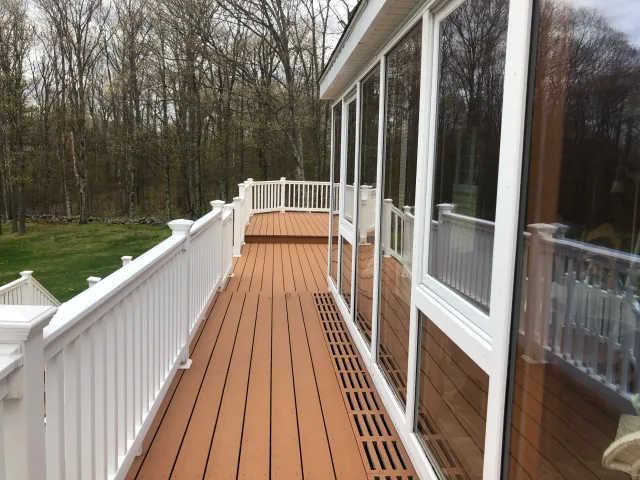 View of a wooden porch with white railings and large glass windows, overlooking a wooded area with leafless trees.