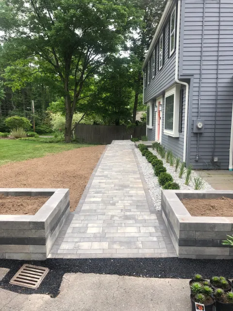 Newly paved walkway with brick pattern leading to the house, bordered by small bushes and white gravel, with a large tree and green lawn on the left and a gray house with multiple windows on the right.
