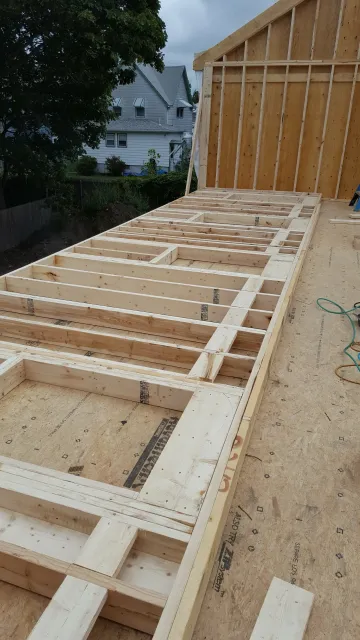 Wood-framed roof deck under construction beside a house, with exposed rafters and plywood sheathing