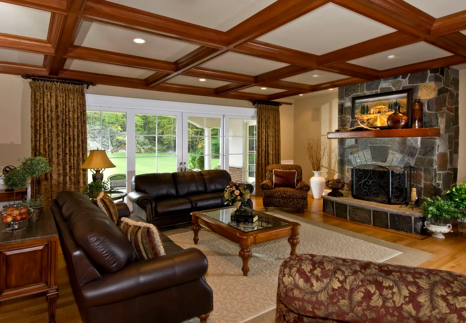 Living room with leather sofas, a stone fireplace, wooden ceiling beams, and large windows showing a green outdoor landscape.