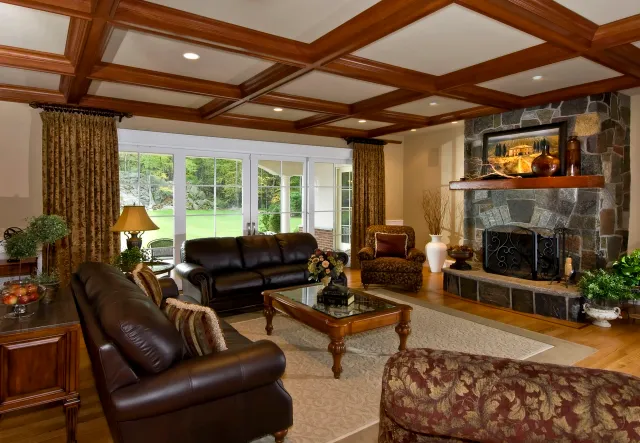 Living room with leather sofas, a stone fireplace, wooden ceiling beams, and large windows showing a green outdoor landscape.
