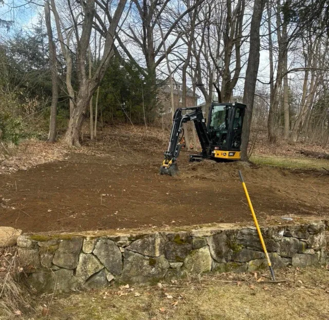 A small excavator working on a dirt clearing in a wooded area, with a stone retaining wall in the foreground and a yellow digging bar leaning against it.