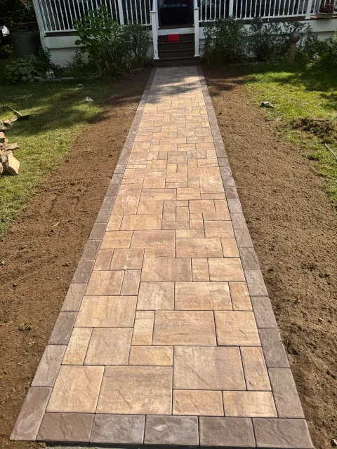 Newly laid rectangular stone pathway leading to a house with steps and a porch, surrounded by grass and garden plants.