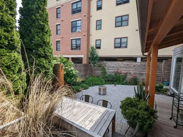 Urban garden patio with outdoor furniture, potted plants, and a fire pit, surrounded by a wooden fence and large buildings in the background.