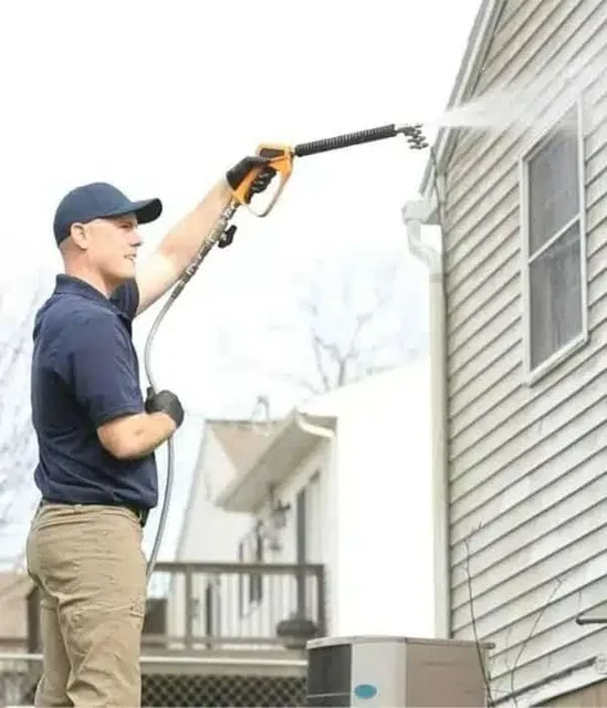 Man power washing a house exterior. He wears a cap and gloves, aiming the pressure washer at the siding to perform exterior cleaning.
