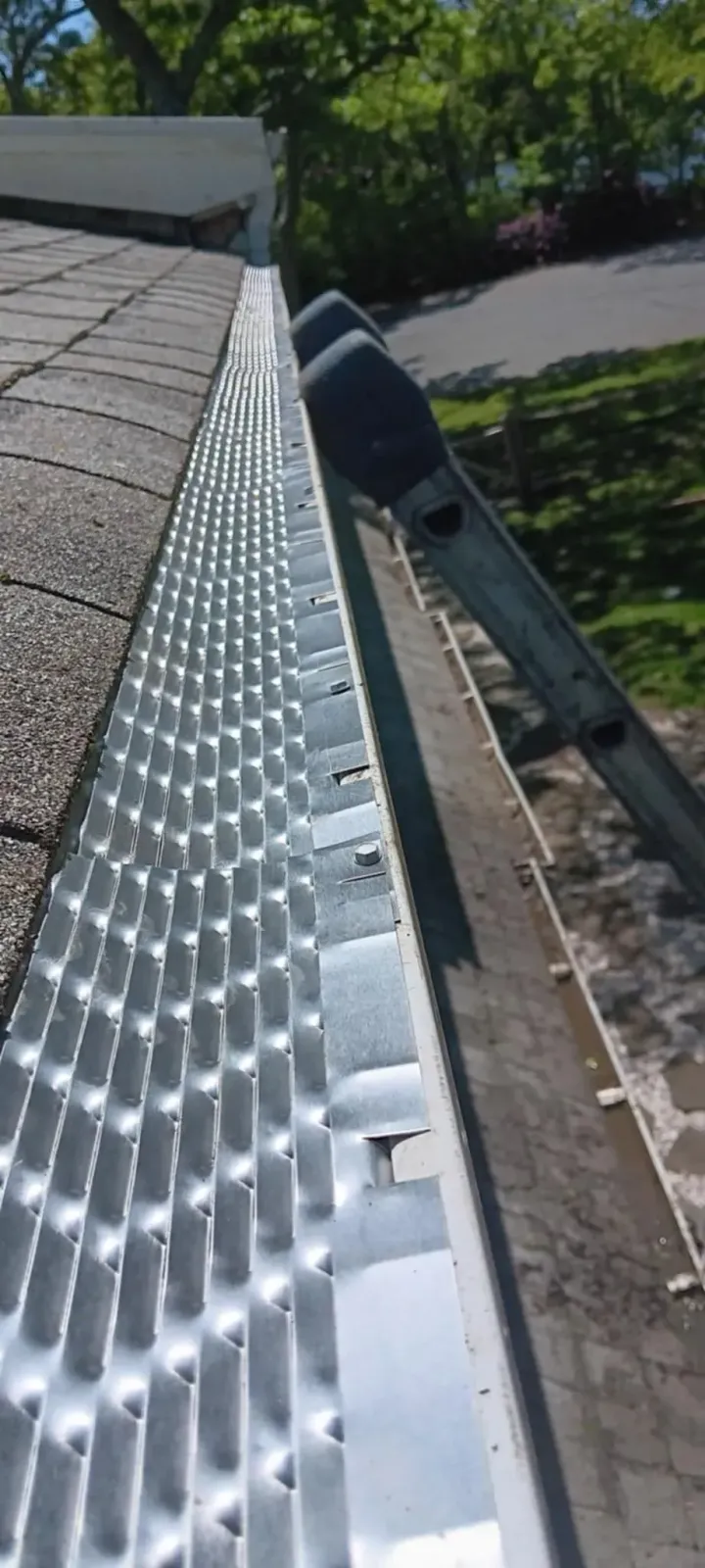 Close-up of a rooftop gutter and shingles with bright sunlight and trees in the background