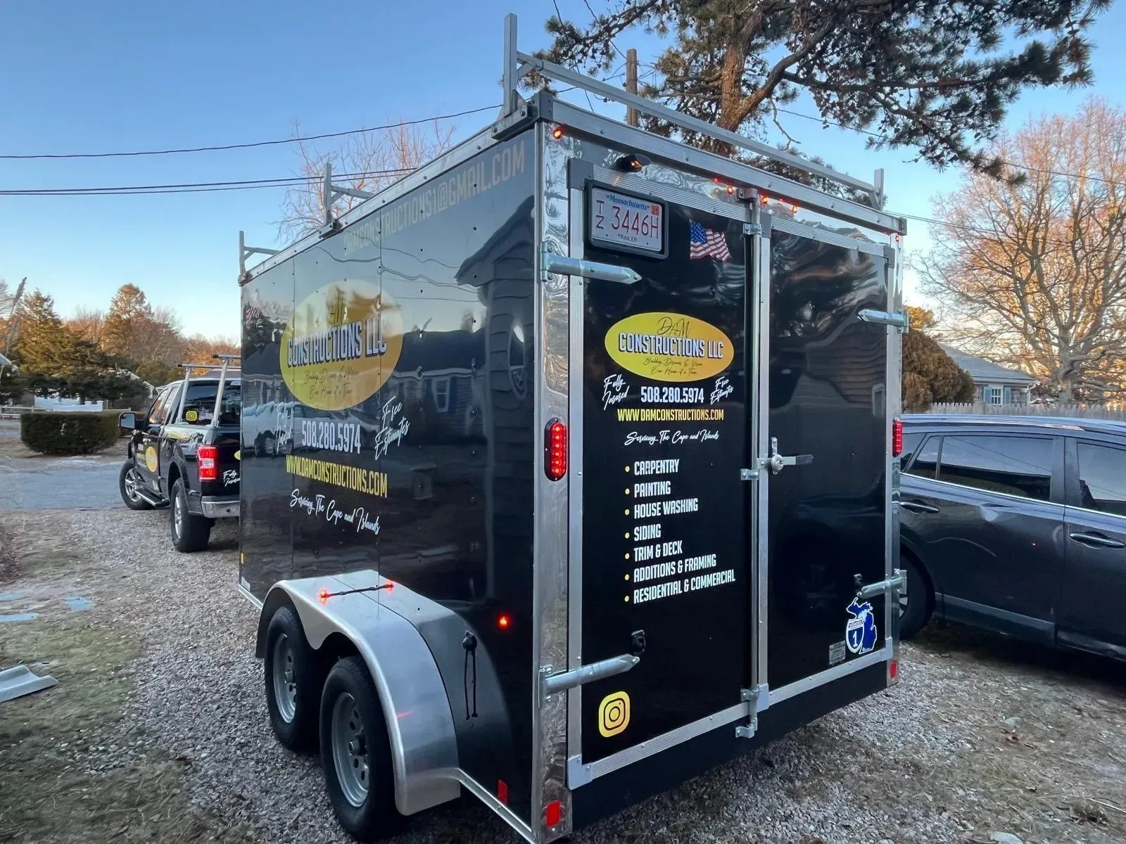 Black trailer with business logo and ladder rack, parked outside, with a truck and car nearby.
