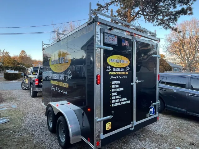 Black trailer with business logo and ladder rack, parked outside, with a truck and car nearby.