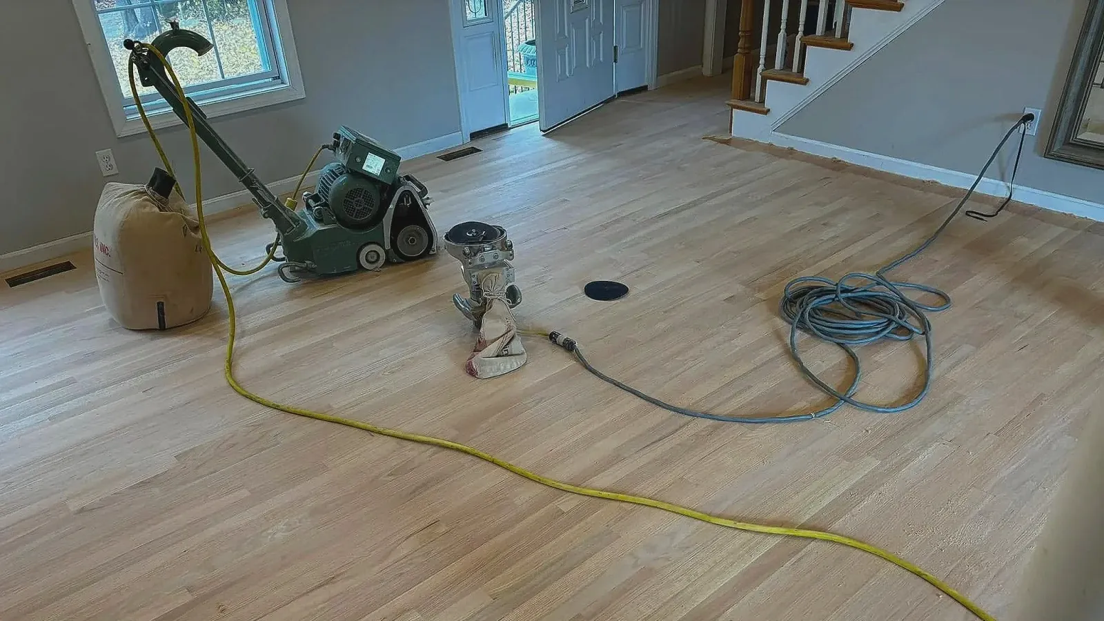A hardwood floor being sanded with rental equipment, including a floor sander, a vacuum, and extension cords, in a living room with a sliding glass door and staircase.