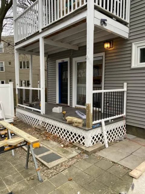 A gray house with a partially constructed deck featuring white railings, lattice skirting, and a pile of lumber.