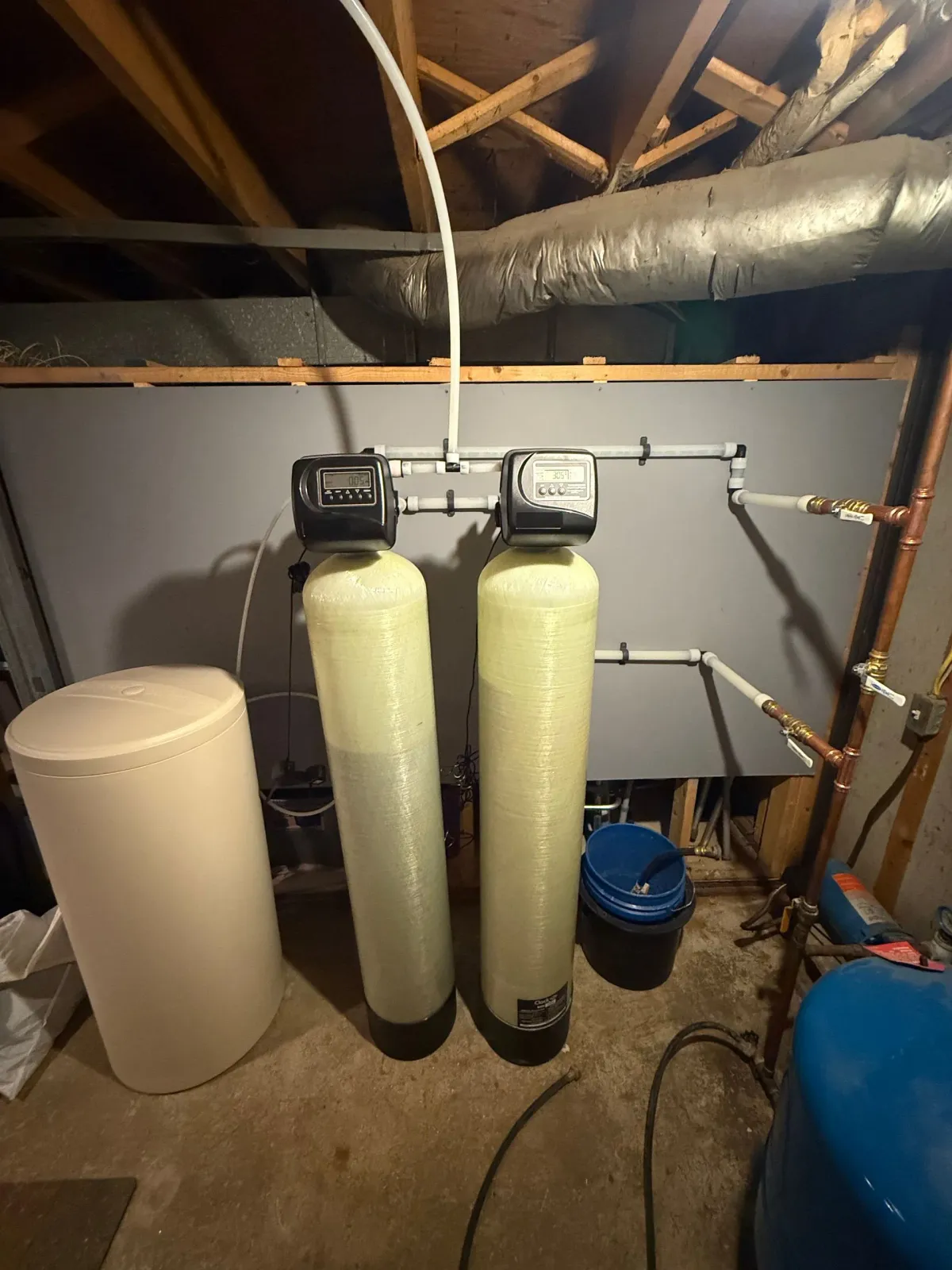 A water treatment system with two tall, beige mineral tanks and control heads, connected by white pipes in a basement.