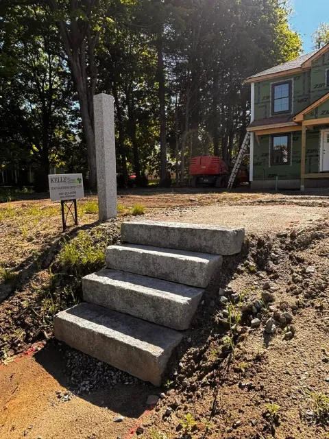 Concrete steps leading up a grassy embankment to a house with trees and a red trailer in the background