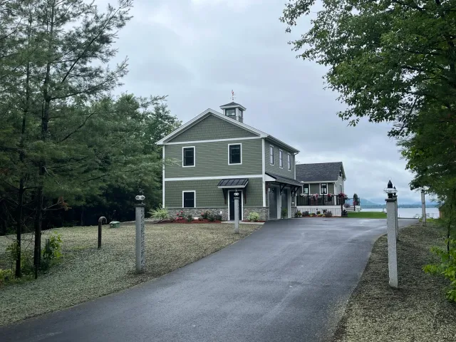 A large gray house with a driveway leading to the waterfront, surrounded by trees and a cloudy sky.