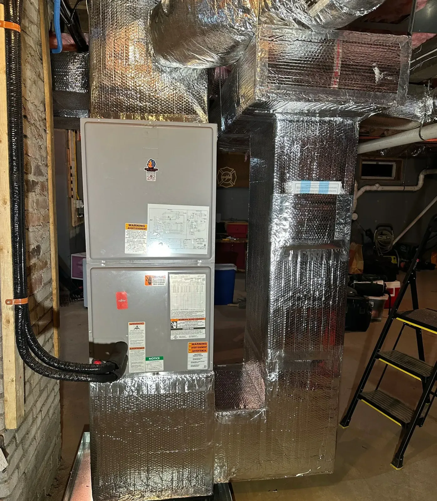 Foil-wrapped HVAC ducts and two gray furnace units in a basement utility room