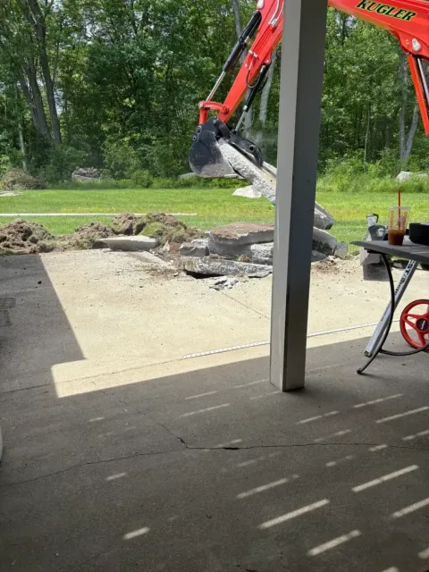 Excavator removing concrete patio slabs in a backyard, with debris and a porch post in the foreground