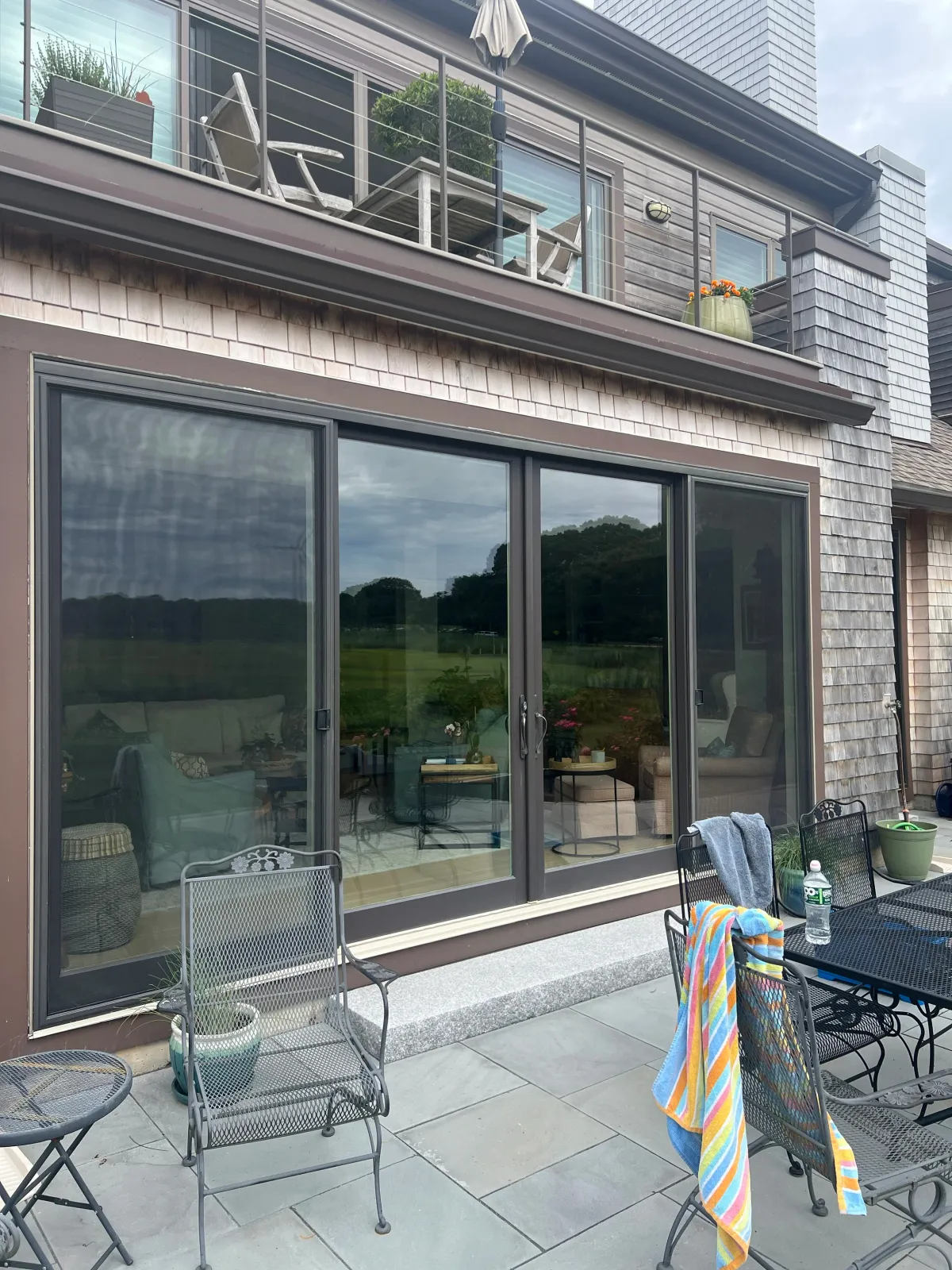 Patio with outdoor furniture and sliding glass doors leading into a house. The patio has chairs, a table, water bottle, and towels. The house has a wooden exterior with a second-floor balcony.
