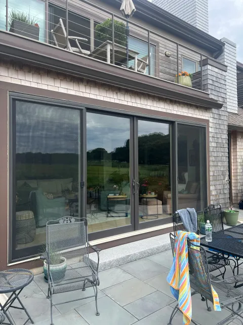 Patio with outdoor furniture and sliding glass doors leading into a house. The patio has chairs, a table, water bottle, and towels. The house has a wooden exterior with a second-floor balcony.
