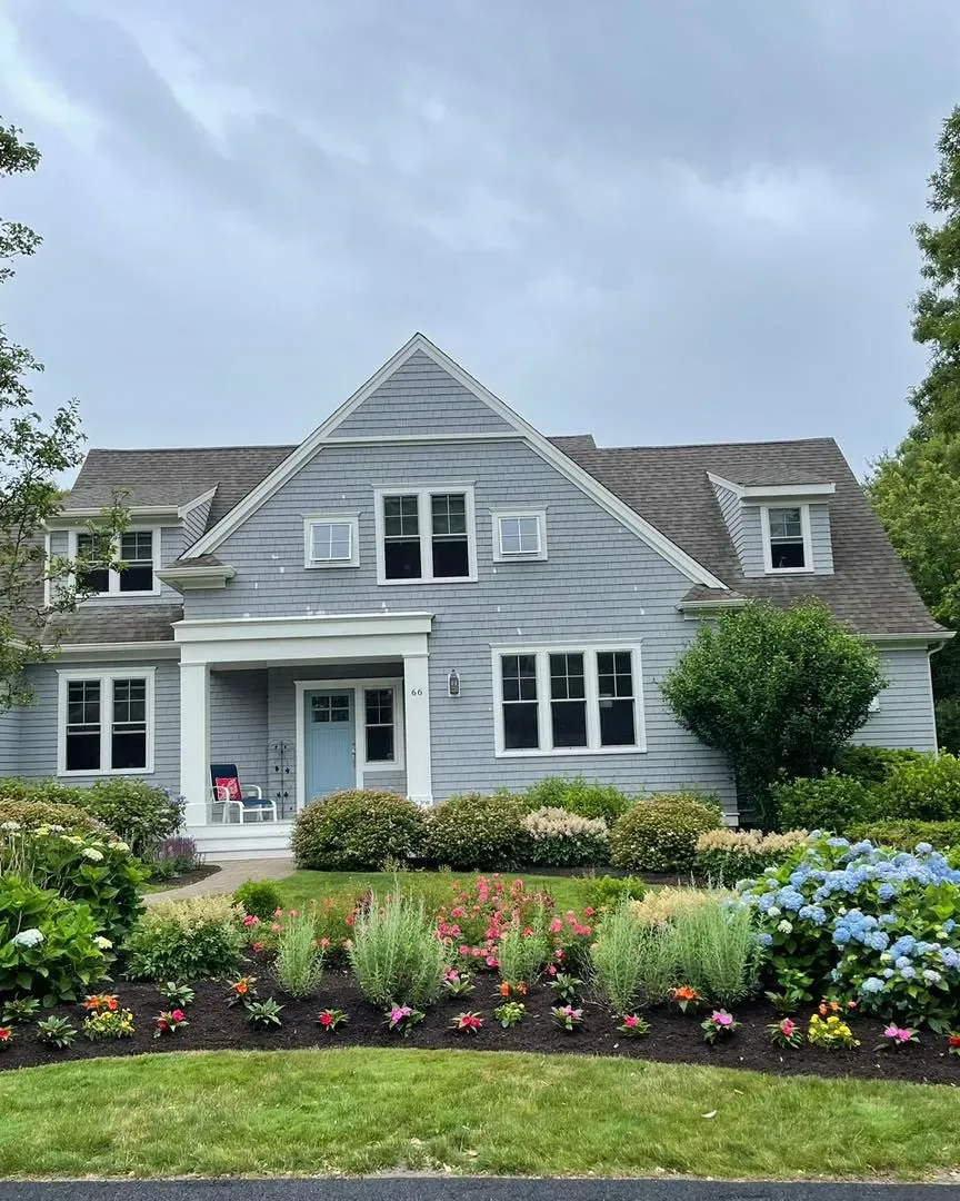 Gray house with blue door and flower garden. Overcast sky.