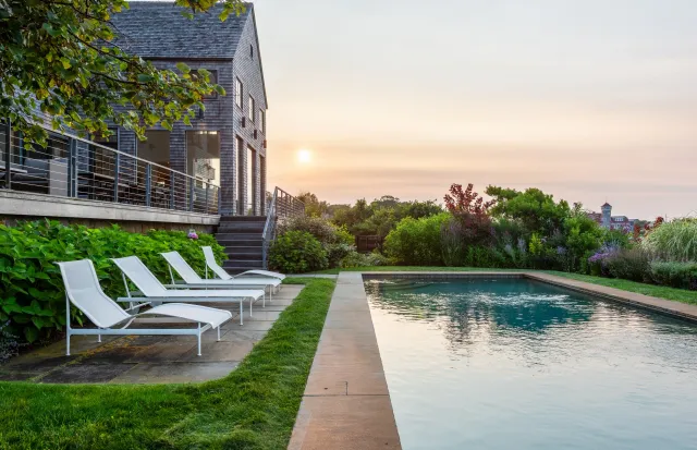 An outdoor swimming pool with four white lounge chairs on a stone patio, surrounded by green bushes and colorful flowering plants, under a sunset sky near a modern house with gray shingle siding.