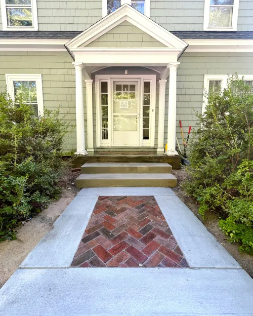 Front porch of a house with brand new concrete steps and a herringbone brick inlay sidewalk leading to the entrance, with bushes on either side.
