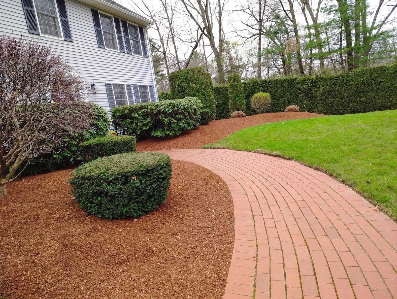 Curved brick walkway beside a house, lined with mulch, shrubs, and green lawn.