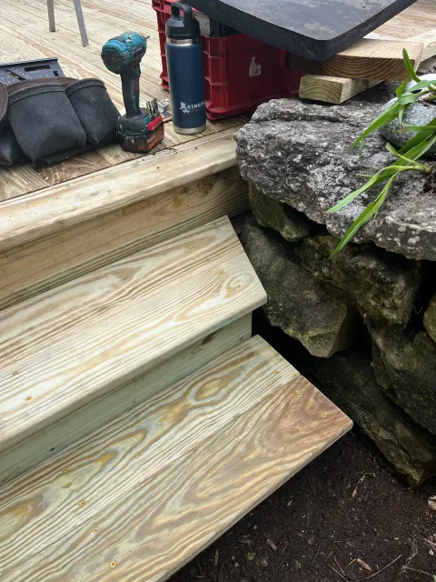 Wooden stairs under construction with tools and supplies on top, next to a stone wall with plants.