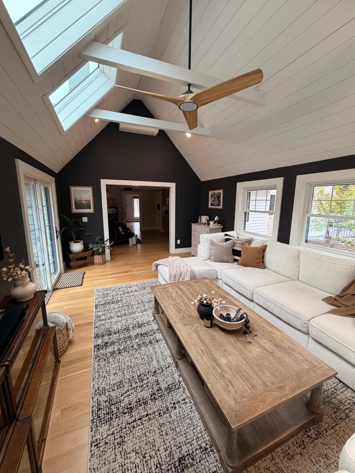 Living room with white sectional sofa, wooden coffee table, area rug, black and white walls, large windows, ceiling fan, framed art, potted plants, and adjacent dining area.