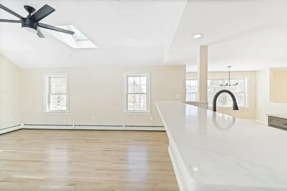 An open-concept room with a light-colored wood floor, white walls, a ceiling fan, and a view into a kitchen area.
