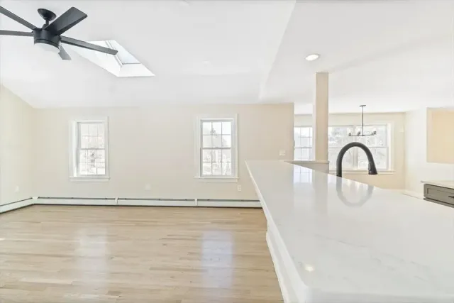 An open-concept room with a light-colored wood floor, white walls, a ceiling fan, and a view into a kitchen area.