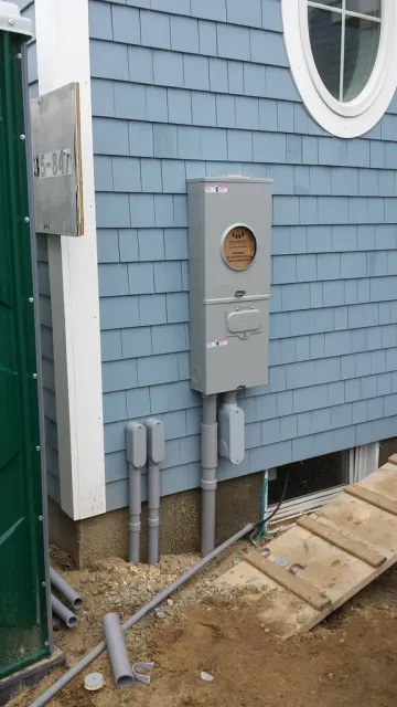 Gray utility meter box with exposed pipes mounted on a blue house exterior near a window and steps