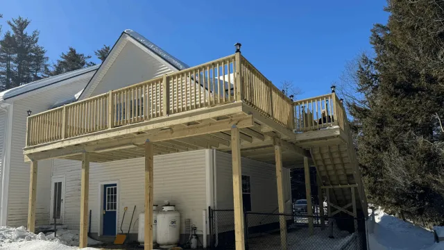 Newly built wooden deck on the second floor of a house, with stairs leading down to the ground, winter snow on the ground, and surrounded by trees.